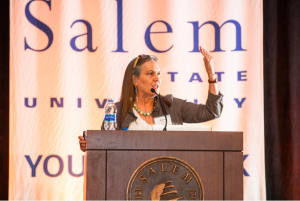 Debby speaking at a podeum in front of sign reading "Salen State University"