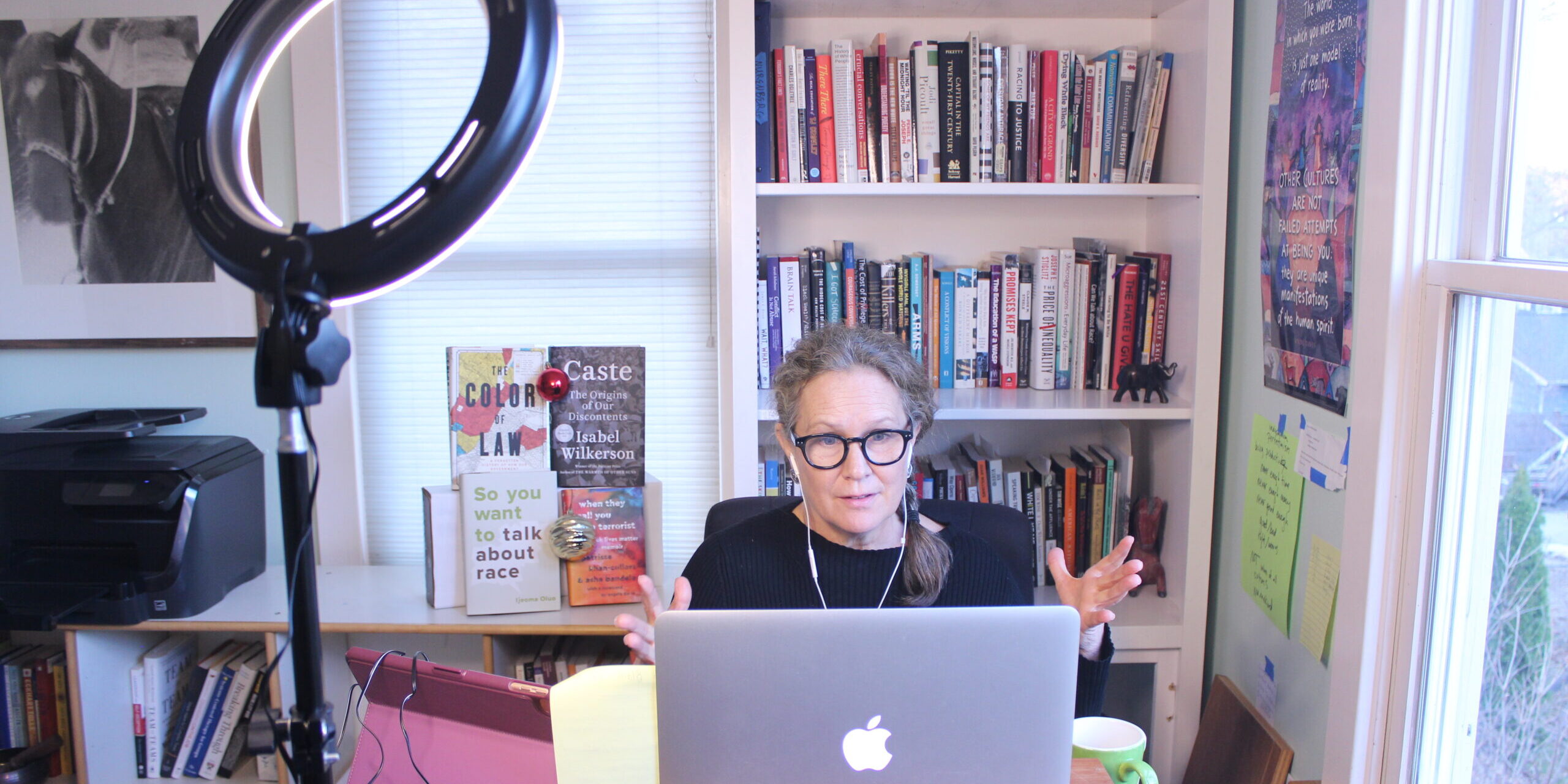 Debby in her office facing her laptop engaged in a presentation. A ring light in the foreground and her bookshelf in the background
